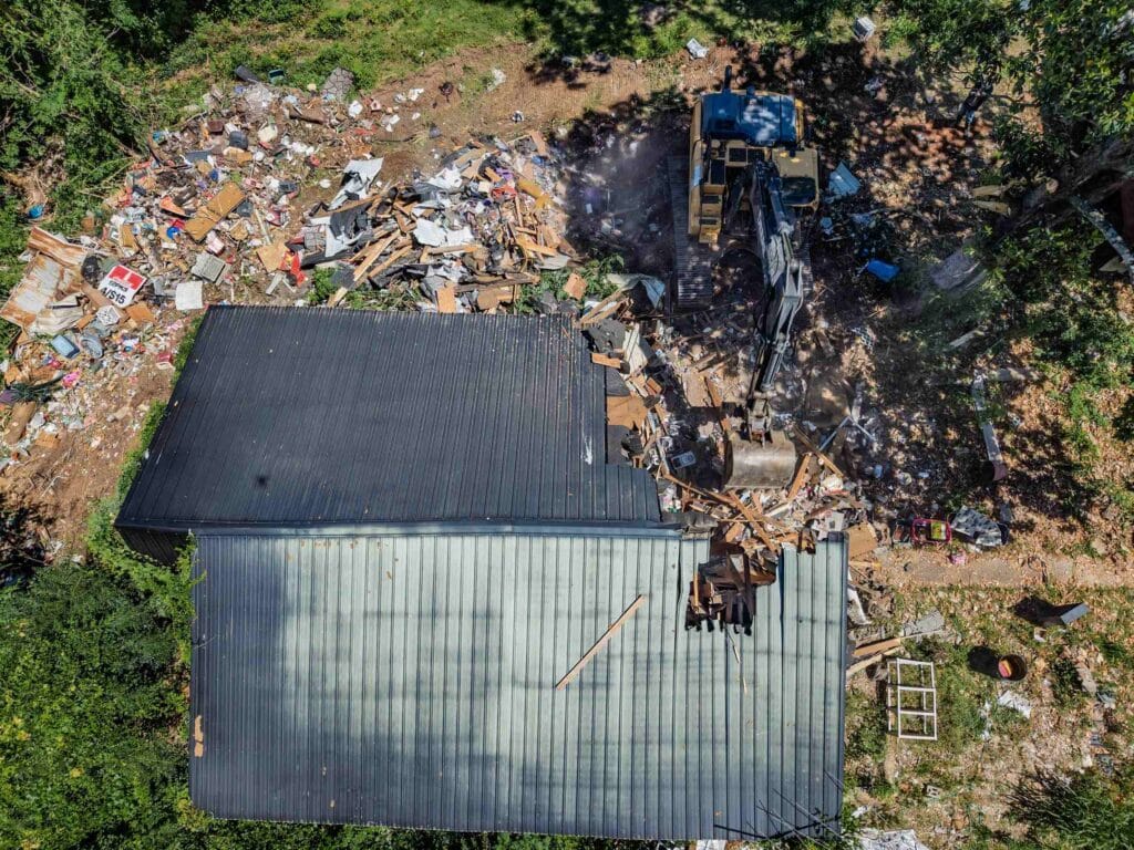 Aerial view of a metal-roofed structure mid-demolition, with a John Deere excavator working beside a large pile of lumber and household debris in Alabama