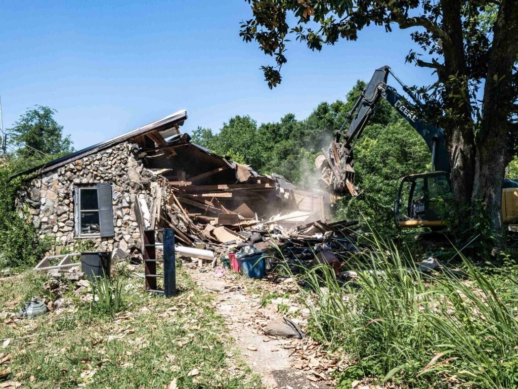 John Deere excavator with a grapple attachment pulling down the roof of a stone-veneer house during a demolition in Alabama
