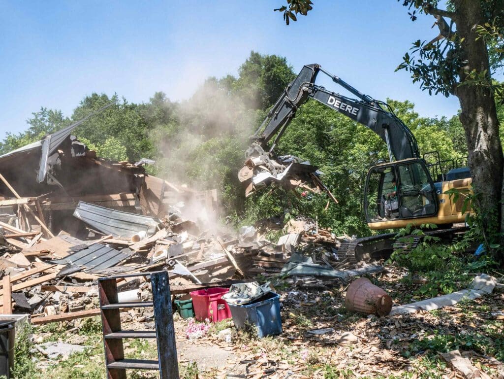 John Deere excavator tearing into a collapsed house as dust rises off the debris pile in Alabama