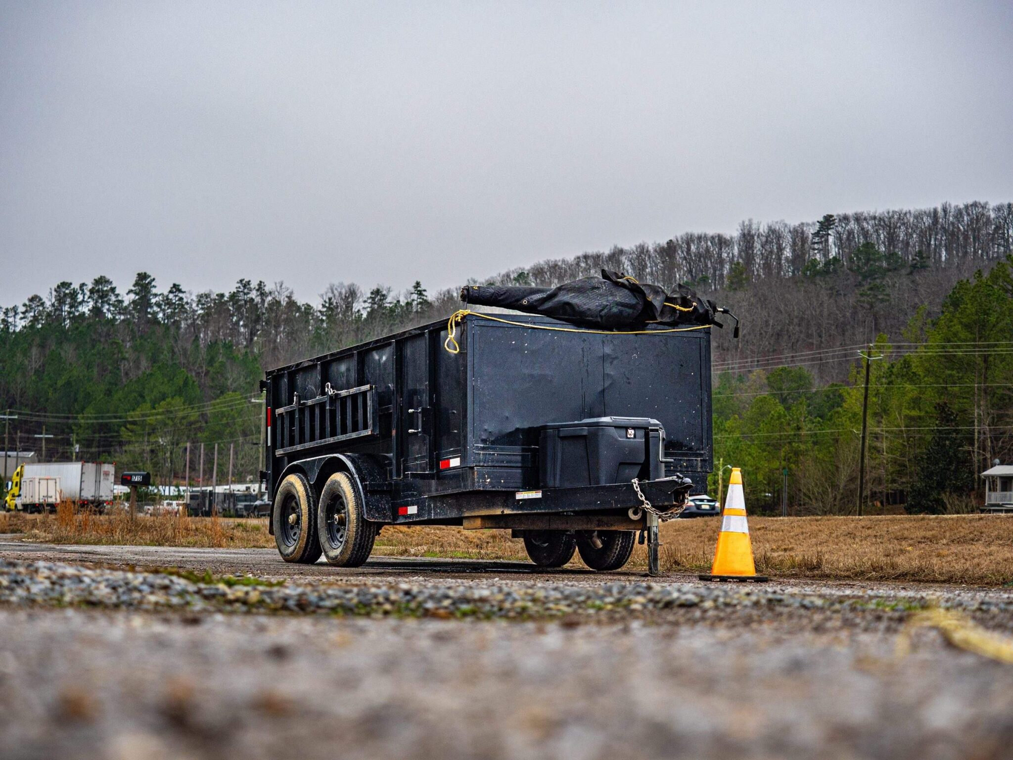 Low-angle view of a dump trailer with a rolled tarp and tongue-mounted toolbox parked on a gravel apron in Alabama