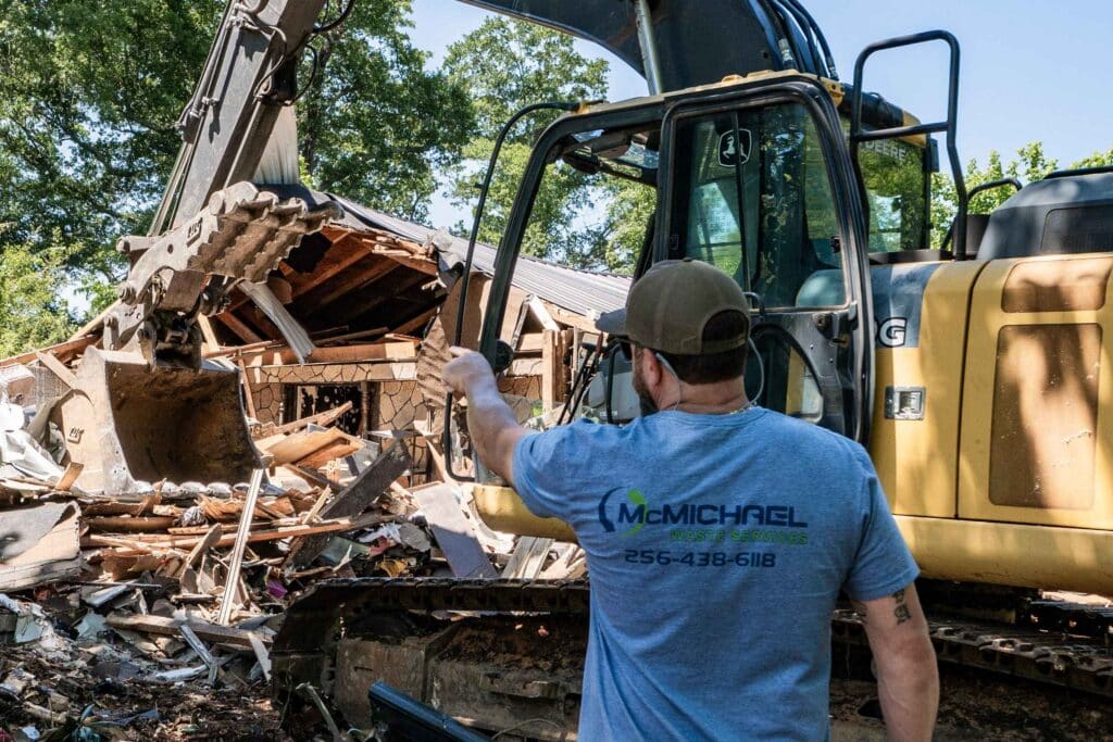 McMichael Waste Services worker in a branded shirt standing at the cab of a John Deere excavator as it tears into a house during a residential demolition in Alabama