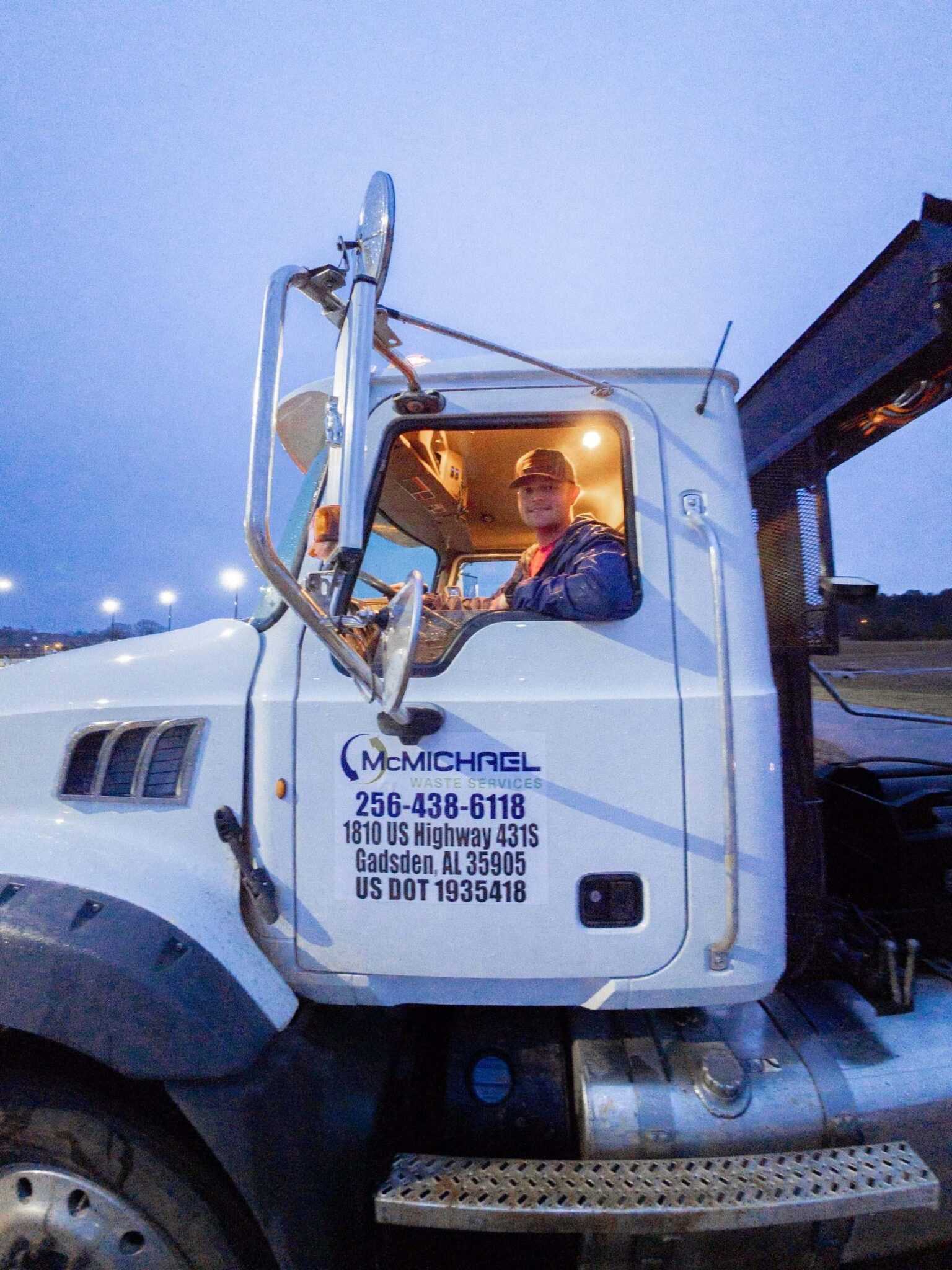 McMichael Waste Services roll-off driver looking out from the cab, showing the full door decal with the Gadsden, Alabama address and US DOT number