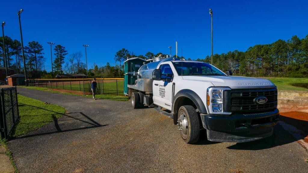 McMichael Waste Services vacuum truck with a polished stainless tank on a service call at an Alabama ball field, the McMichael door decal and 256-438-6118 phone number visible on the cab door