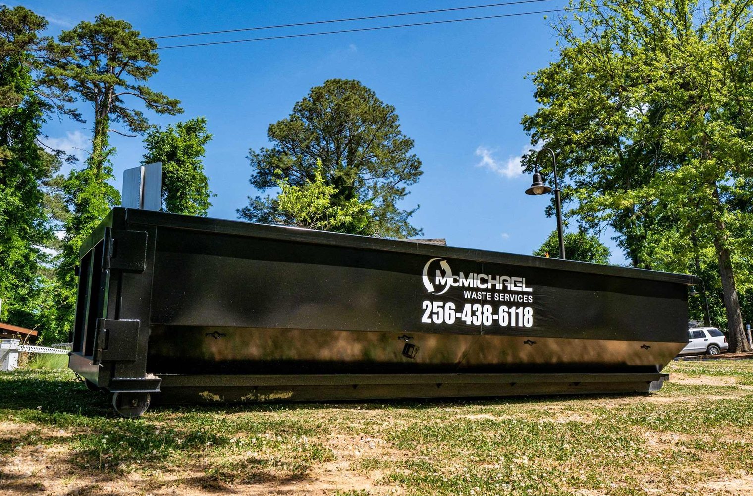 McMichael Waste Services roll-off dumpster with 256-438-6118 set on a grassy lot under trees
